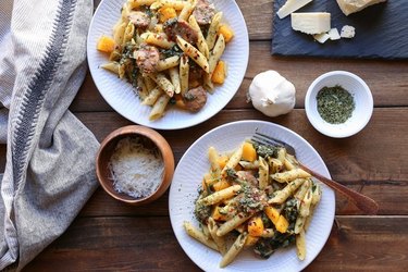 Overhead view of two bowls of penne with sausage and squash, on a wooden table surrounded by a kitchen towel and the dish's ingredients.