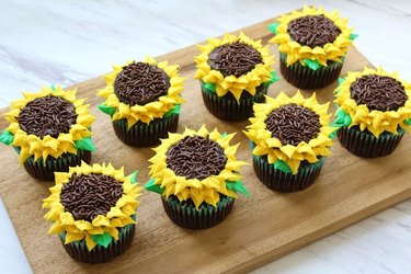 Eight cupcakes, decorated to look like sunflowers, arranged on a wooden cutting board on a marble counter top.