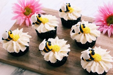 Cupcakes decorated with piped flowers and bumble bees, displayed on a cutting board with a pair of pink flower blossoms.