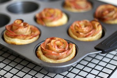 Soft-focus image of "blooming flower" apple pies in a muffin pan, sitting on a wire cooling rack.