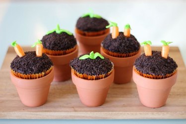 A set of six cupcakes in small terra cotta pots, with tops decorated to resemble baby plants in soil.