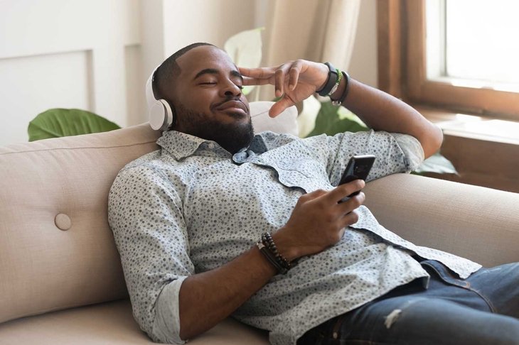 African calm guy hold smart phone fell asleep lying on couch listens music on headphones enjoy song or meditation audio course feels serenity inner harmony and balance, no stress mood, leisure concept