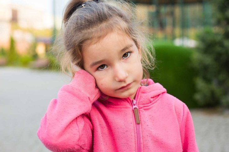 A little girl in a pink hoodie with a sad and tearful face is holding her ear. Ear pain, otitis media, swelling of the cheek, gums, toothache, children's surgery, otolaryngology. Children's medicine